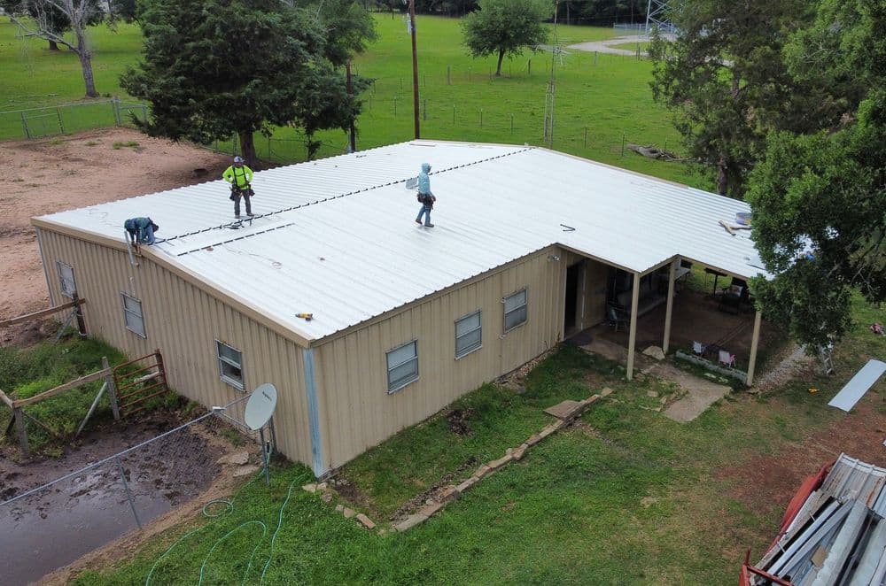 Workers installing a metal roof on a rural building surrounded by green fields.
