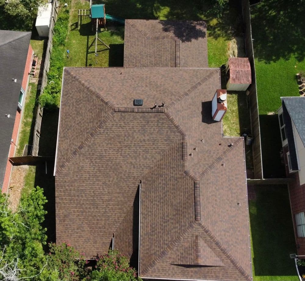 Aerial view of a residential roof with dark shingles and green lawn in background.