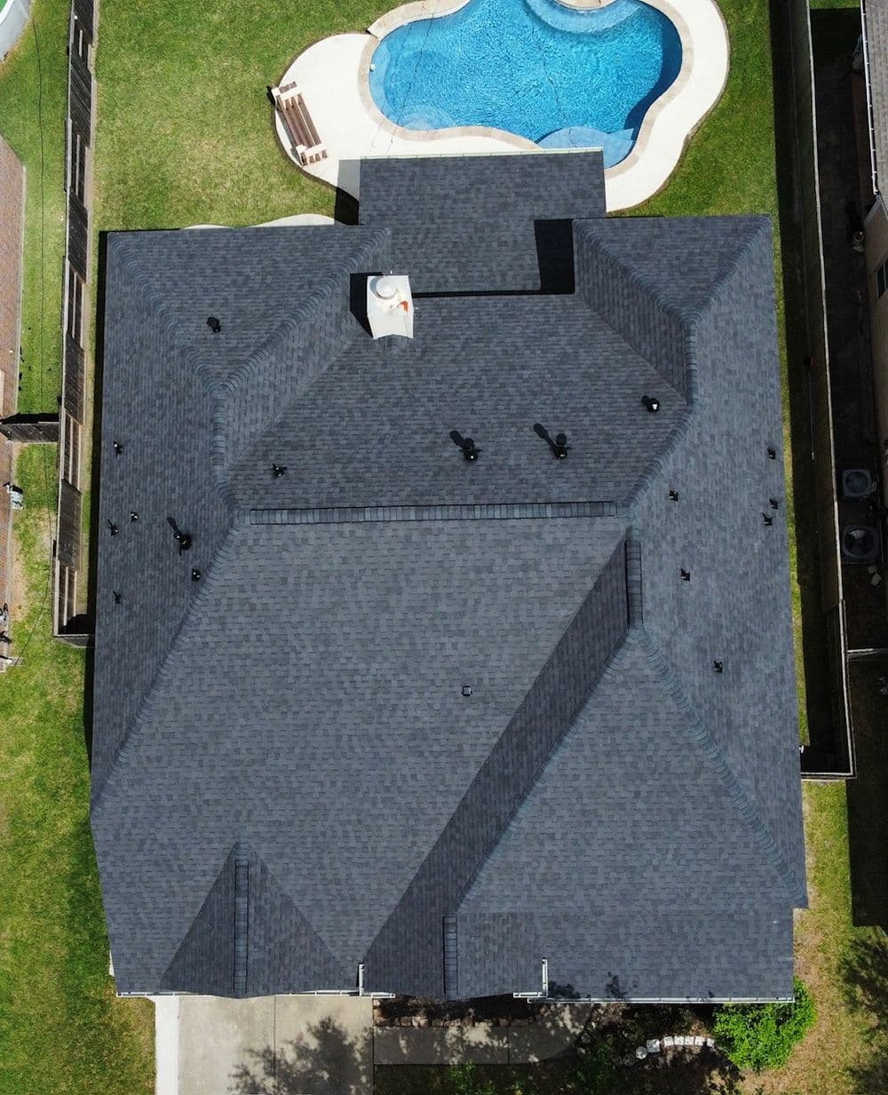 Aerial view of a dark shingle roof with a swimming pool in the background.