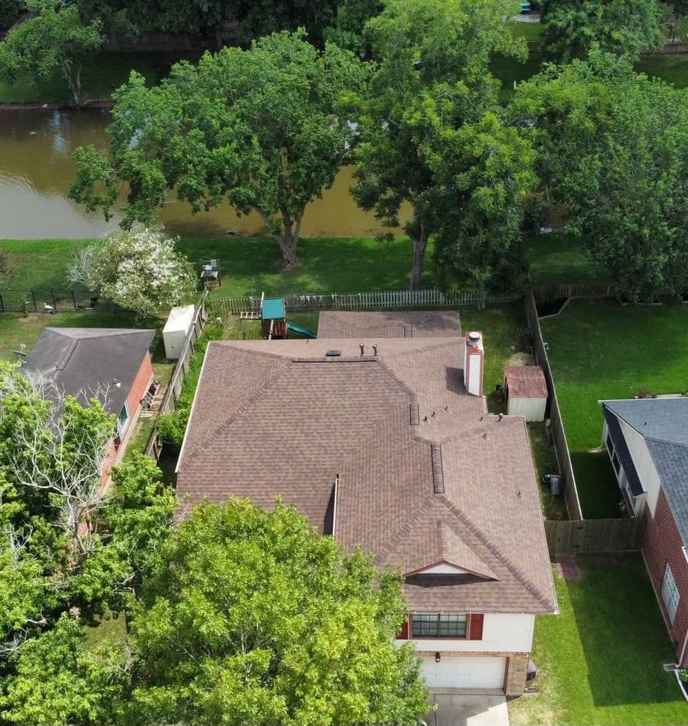 Aerial view of a residential home with a brown shingle roof near a river and green trees.