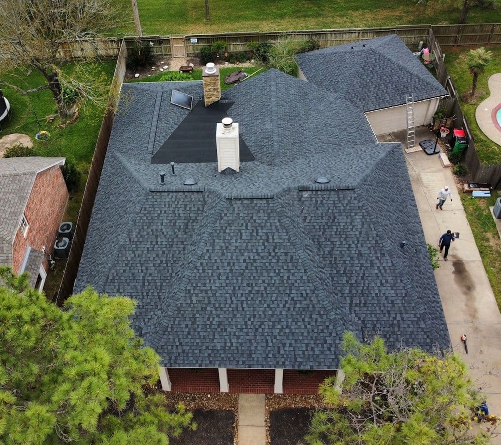 Aerial view of a dark shingle roof on a home with a chimney and nearby landscaping.