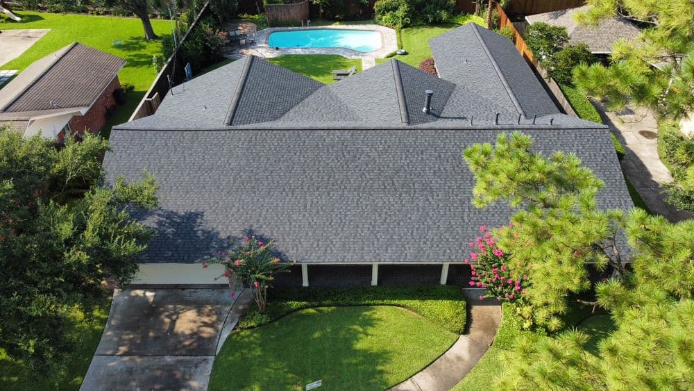 Aerial view of a home with a dark shingle roof, surrounded by greenery and a pool.