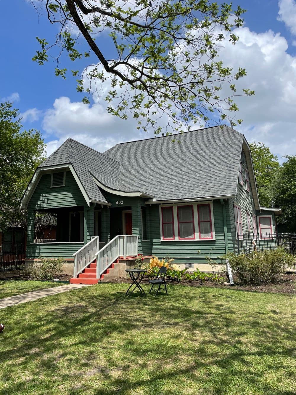 Green Victorian-style house with a steep roof and red accents, surrounded by a lush yard.